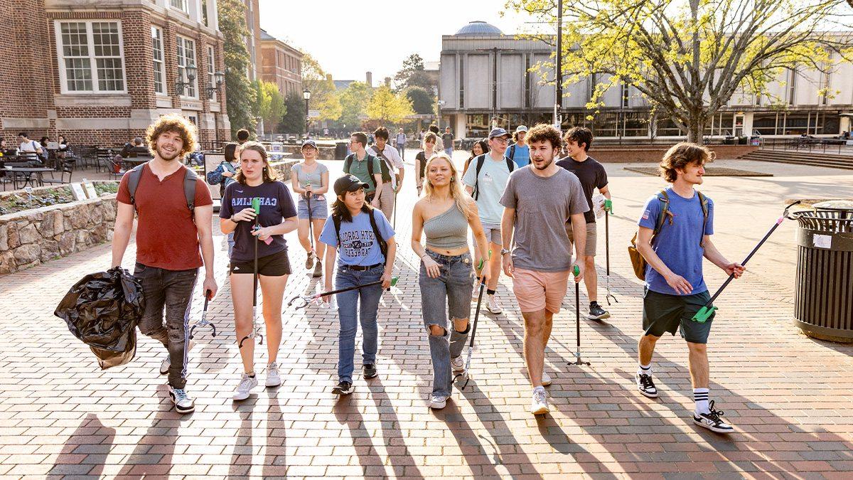 Students walking along brick pathways on the campus of UNC-Chapel Hill holding trasha pickers and trash bags.